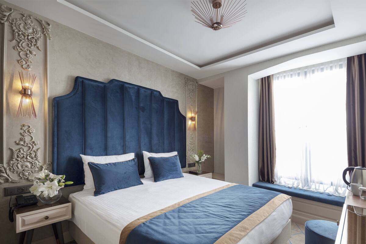 Una habitación de hotel moderna con una cama grande, una mesita de noche con lámpara, una ventana con cortinas y un panel decorativo en la pared.