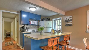 A kitchen with blue cabinets, a white refrigerator, a microwave, and a coffee station. There are three wooden bar stools and a window with blinds.