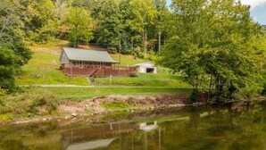 A cabin with a porch and a small shed, surrounded by a grassy area and a body of water.