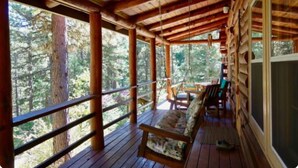 Una terraza de madera con una silla mecedora, un banco y una mesa, rodeada de árboles altos.
