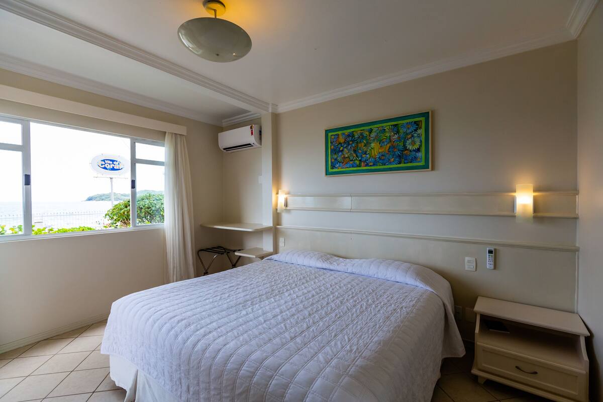 Habitación de hotel con una cama grande, una ventana con cortinas, un aire acondicionado de pared, un estante con un cuadro decorativo y una mesita de noche con lámpara.