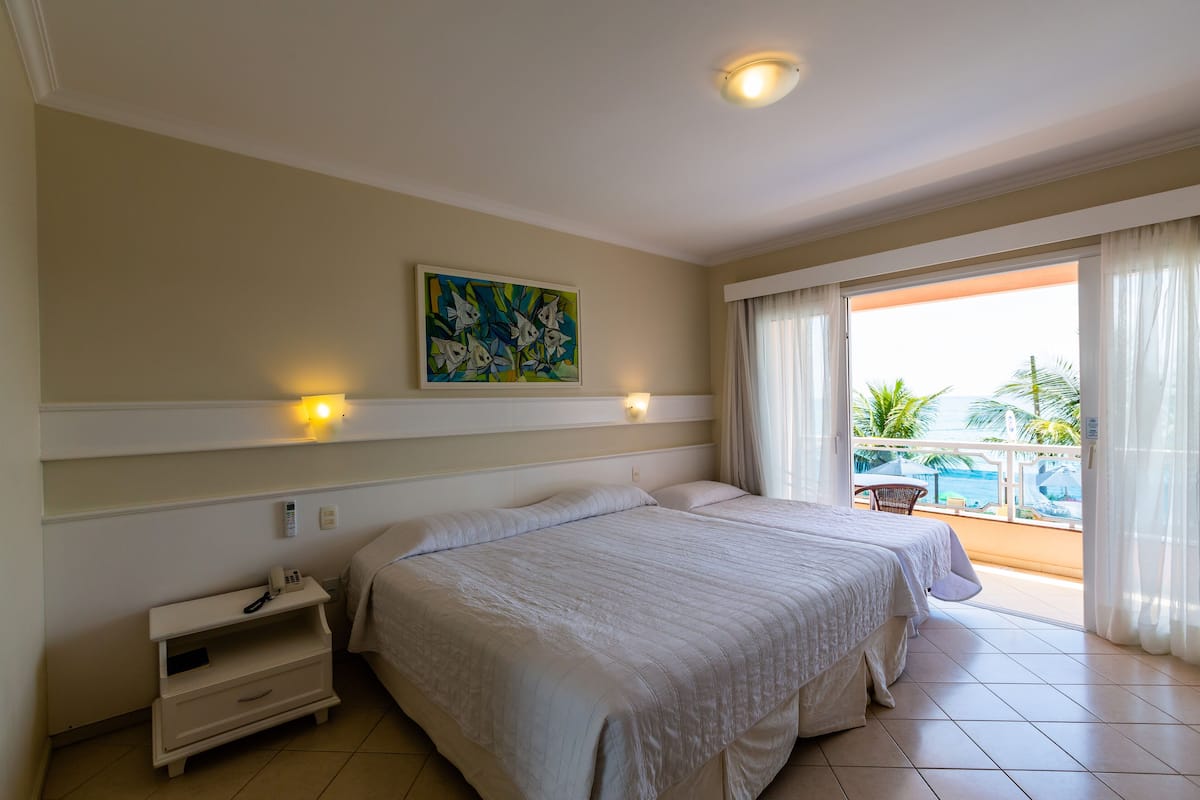Habitación de hotel con dos camas, una mesita de noche, un cuadro en la pared y un balcón con vistas al océano.