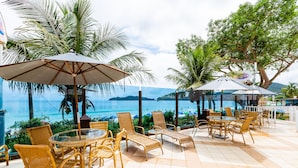 Een terras buiten met rieten meubels en parasols, met uitzicht op een strand en de oceaan.