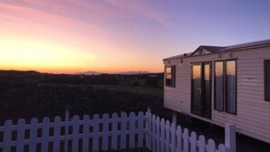 Una casa móvil con fachada blanca y ventanas amplias, rodeada por una cerca de piquetes blancos, al atardecer.