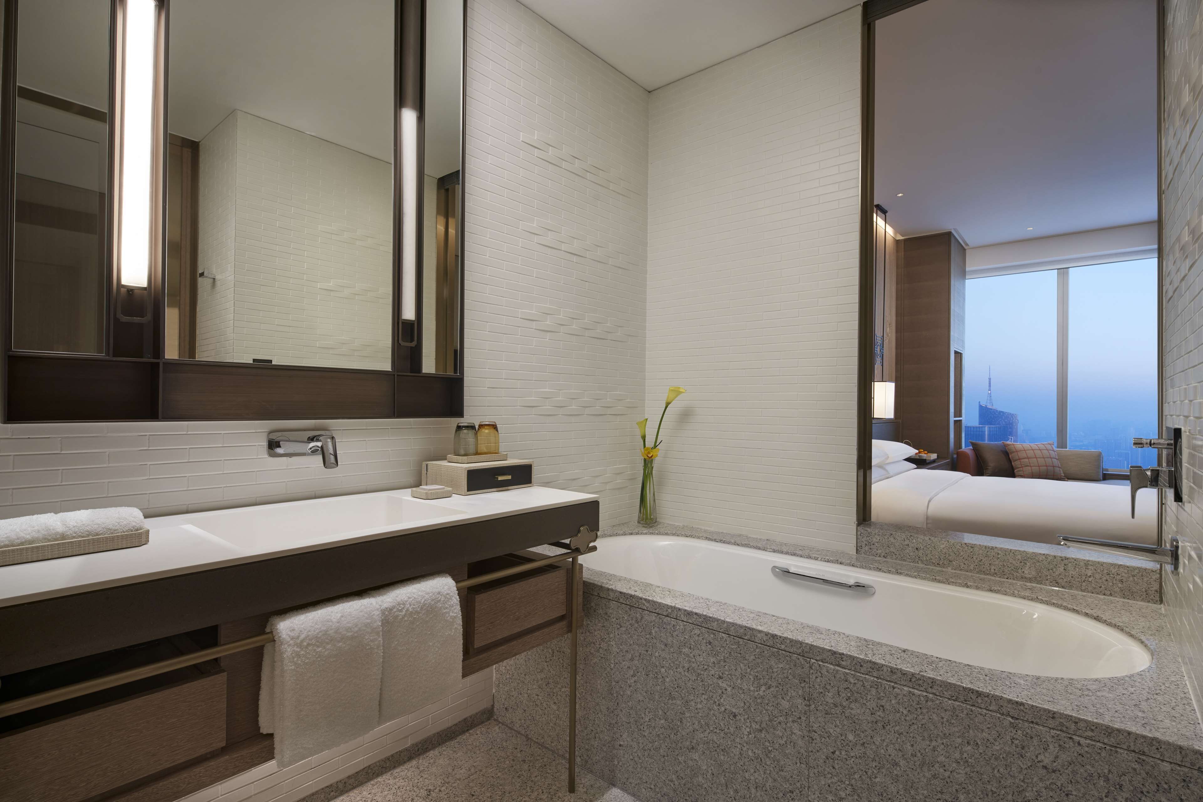 Un baño moderno con un espejo grande, dos lavamanos, una bañera y vistas a un dormitorio con cama y ventana.