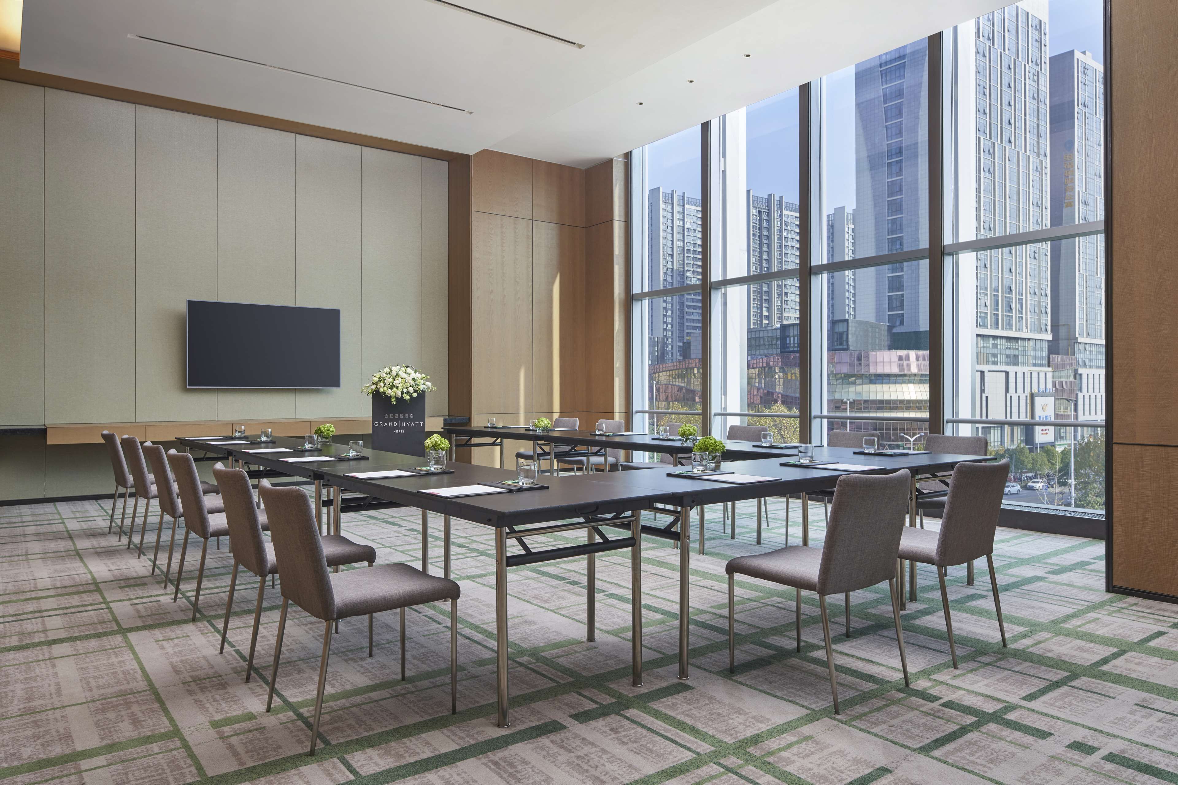 A conference room with a large table, chairs, a flat-screen TV, and a view of tall buildings through large windows.