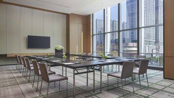 A conference room with a large table, chairs, a flat-screen TV, and a view of tall buildings through large windows.