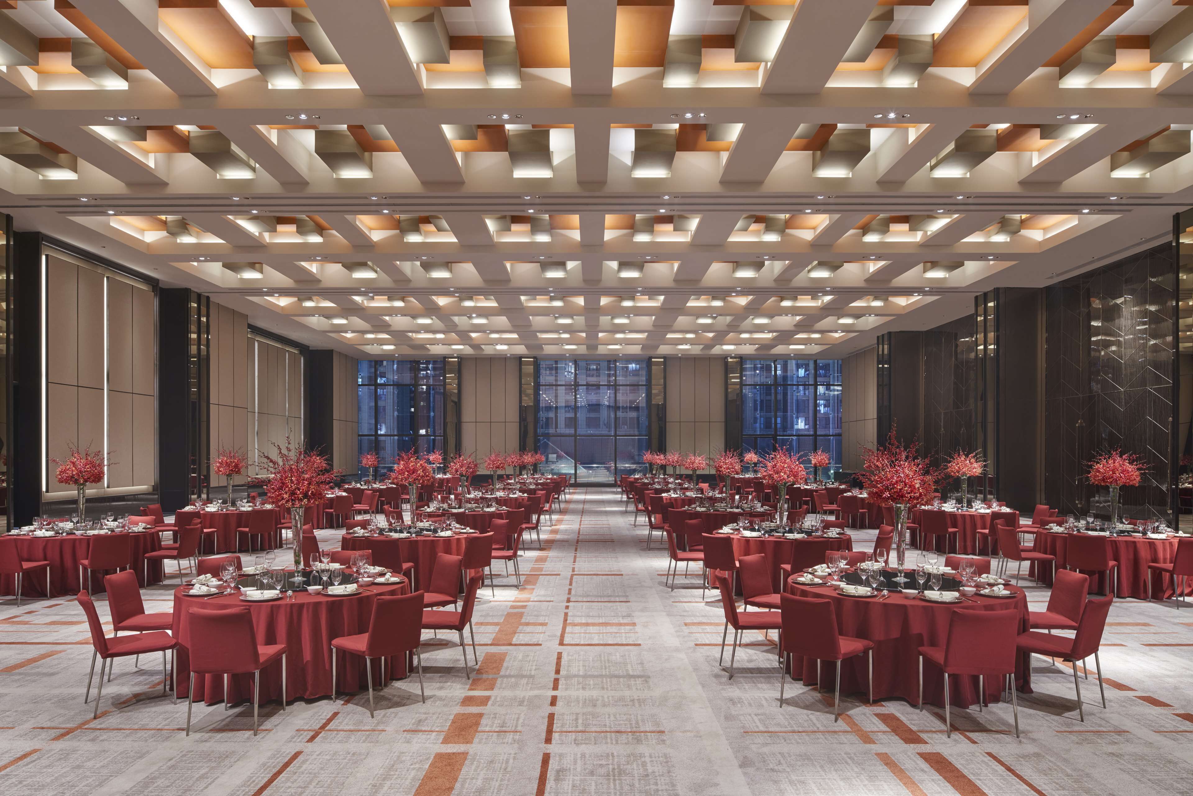 A spacious banquet hall with multiple round tables set for dining, featuring a modern ceiling design and large windows providing a view of the cityscape.