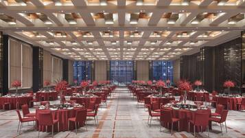 A spacious banquet hall with multiple round tables set for dining, featuring a modern ceiling design and large windows providing a view of the cityscape.