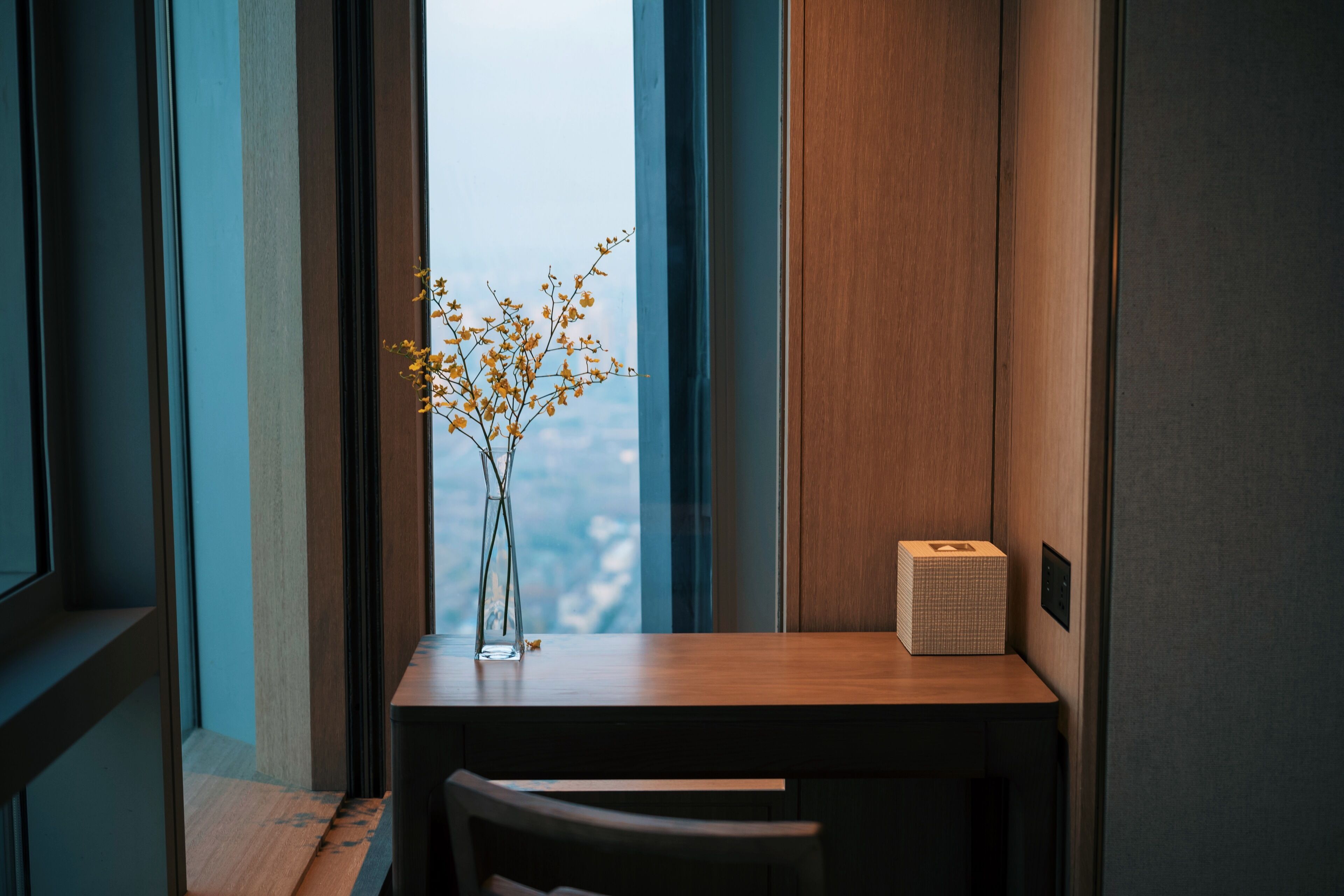 Een moderne kamer met een houten bureau, een vaas met gele bloemen en een uitzicht op de stad door grote ramen.
