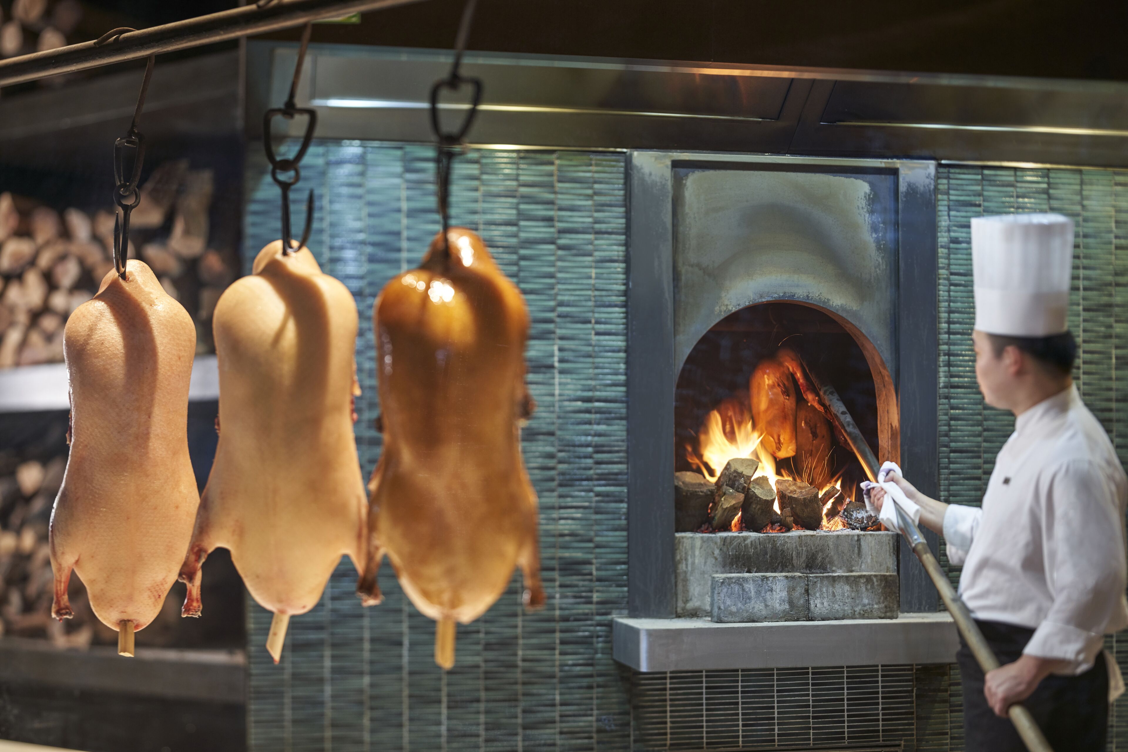 A chef preparing roasted meats in a professional kitchen with hanging racks and a wood-fired oven.