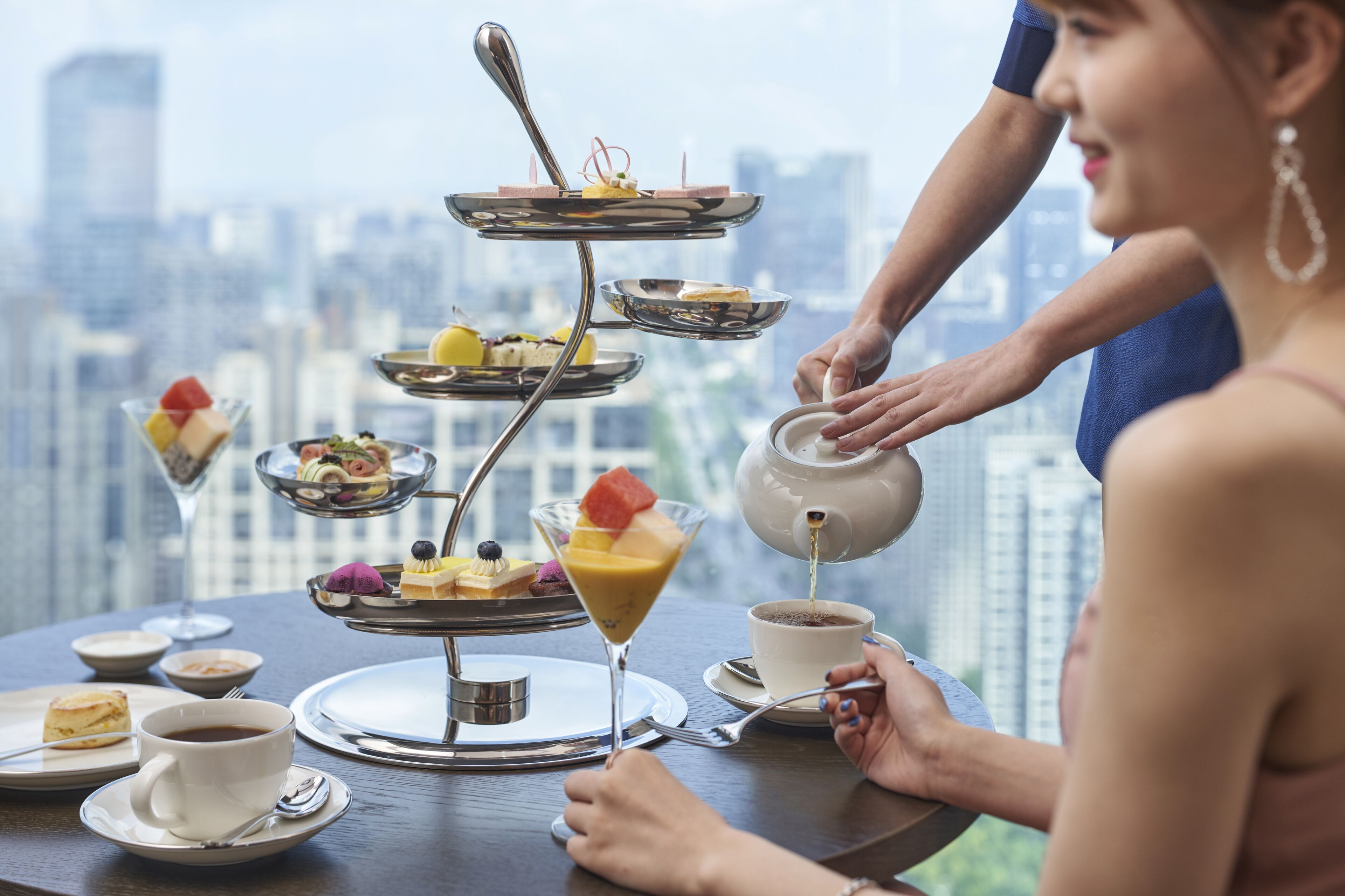 Una bandeja de postres de varios niveles con dulces y frutas, una taza de café y una tetera sirviendo té, sobre una mesa con vista a la ciudad de fondo.