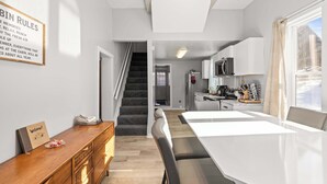 Eine moderne Küche mit weißen Schränken, einer Mikrowelle und einer Kaffeemaschine. Es gibt eine hölzerne Bank und einen weißen Esstisch mit Stühlen. Eine Treppe führt in einen anderen Raum.