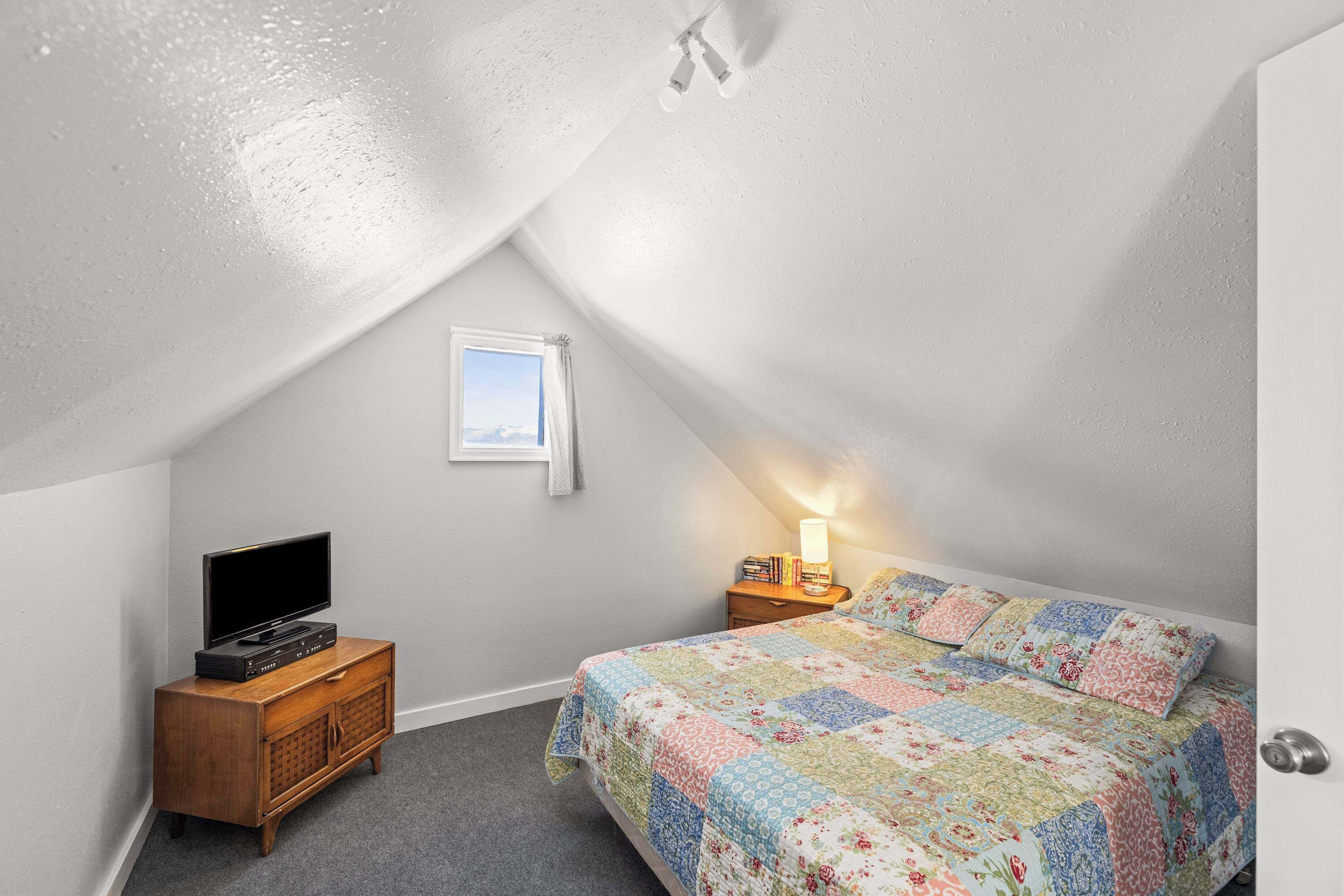 A bedroom with a sloped ceiling, a flat-screen TV on a wooden dresser, a bedside table with a lamp, and a colorful quilt on the bed.