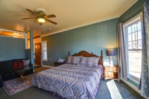 Quarto com cama, ventoinha no teto, mesa de cabeceira, luminária, janela com cortinas e pequena mesa com cadeira.