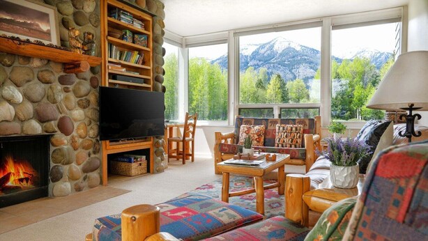 Ein gemütliches Wohnzimmer mit Kamin aus Stein, einem Fernseher, einem Bücherregal und Blick auf Berge durch große Fenster.