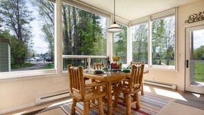 Ein Essbereich mit Holztisch und Stühlen, großen Fenstern mit Blick auf Bäume und ein Haus, sowie einer hängenden Leuchte.