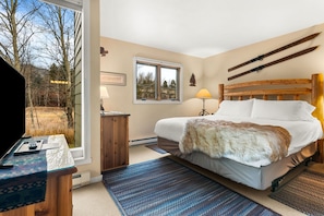 Une chambre à coucher avec un lit, des lampes de chevet, une tête de lit en bois, un bureau avec une lampe et une vue sur l’extérieur.