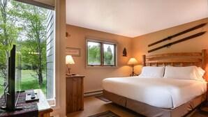 Ein Schlafzimmer mit einem großen Bett, zwei Nachttischlampen, einem Holz-Kopfteil, einem Fenster mit Blick auf Bäume und einem Schreibtisch mit Computer und Lampe.