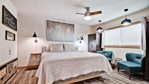 Een slaapkamer met een bed, een plafondventilator, een raam met gordijnen en een kunstwerk aan de muur.
