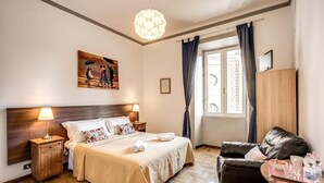 Une chambre d’hôtel comprenant un lit, une tête de lit en bois, une table de chevet, un bureau avec une chaise, une lampe, un tableau et une fenêtre avec des rideaux.