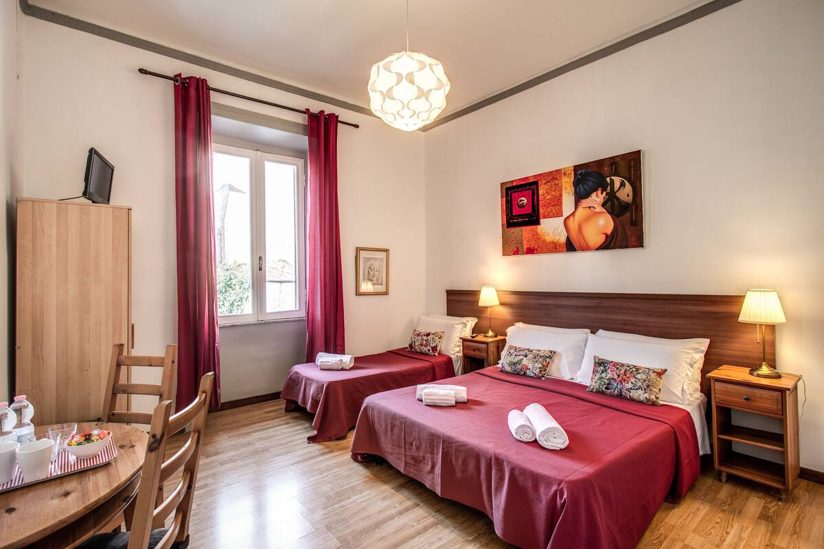 Habitación de hotel con dos camas individuales, un escritorio de madera con silla, una mesa de comedor y un ventanal con cortinas rojas.