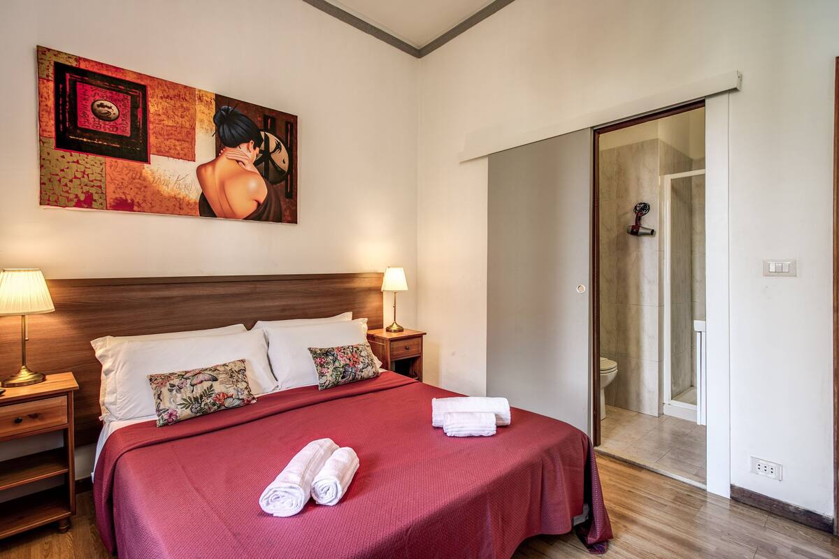 Una habitación de hotel con una cama grande, dos mesitas de noche con lámparas, un cuadro en la pared y una puerta corredera que da a un baño.