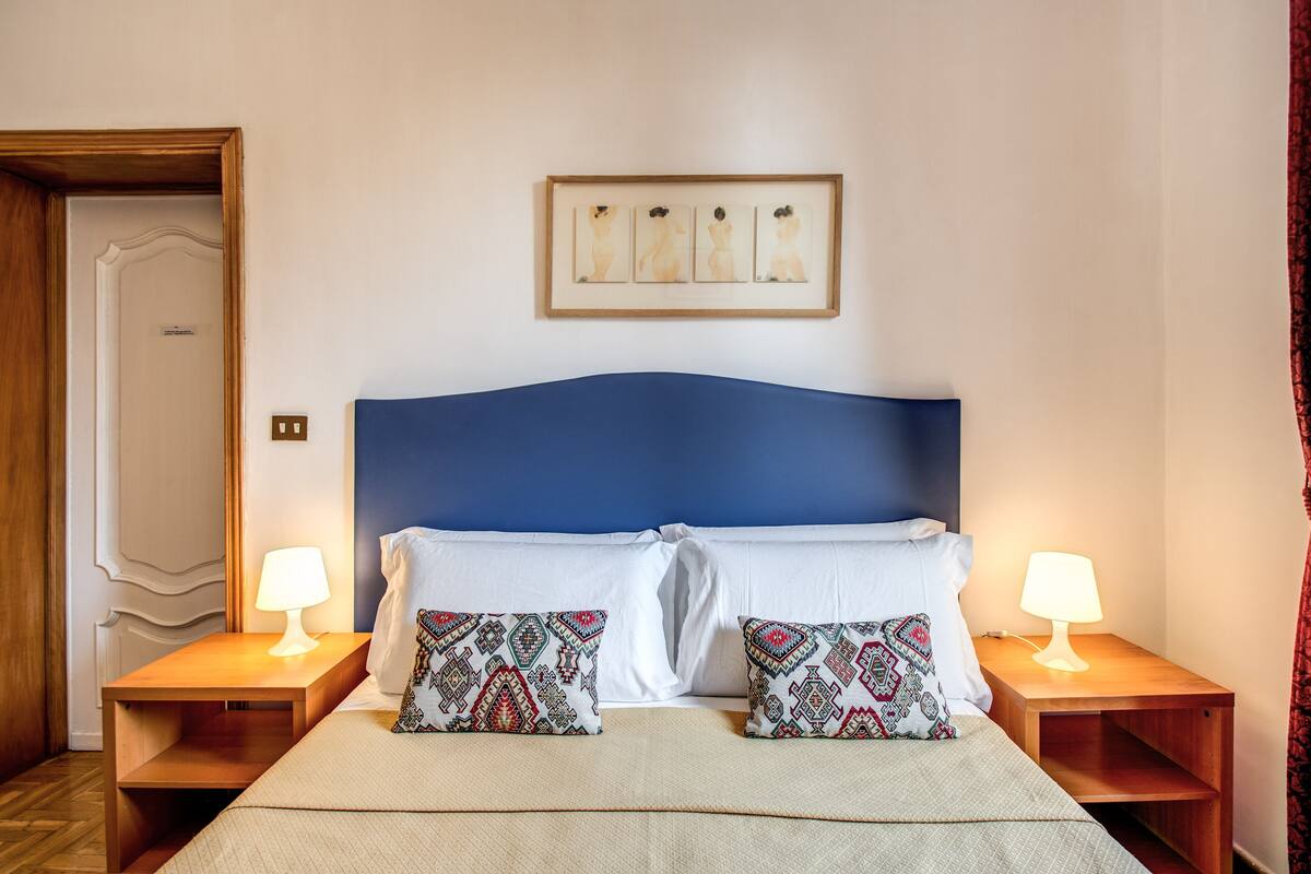 Una habitación de hotel con una cama, dos mesitas de noche con lámparas, almohadas estampadas y una obra enmarcada sobre la cama.