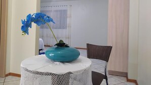 Ein Zimmer mit einem runden Tisch, einem Stuhl, einer Vase mit blauen Blumen und einem Fenster mit Vorhängen.