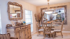 Un coin repas avec une table ronde, des chaises et un lustre. Un grand miroir est placé au-dessus de la table, et un meuble contient des objets décoratifs.