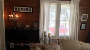 A cozy living room with a wooden wall, a dark wooden cabinet, a beige sofa with a decorative pillow, a chandelier, and a window with sheer curtains. - Romantic Slipper Tub (Carmine)
