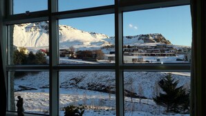 A view through a window with multiple panes. The top pane shows a snow-covered mountain with a clear sky. The middle pane shows a residential area with houses and a road. The bottom pane shows a snowy landscape with trees and a fence. - Ársalir Guesthouse (Vik I Myrdal)