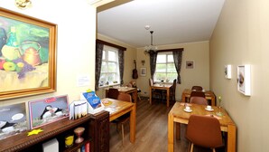 A dining area with wooden tables and chairs, a painting on the wall, and a window with curtains. - Ársalir Guesthouse (Vik I Myrdal)