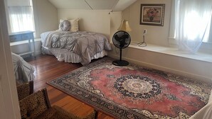 A bedroom with a bed, a standing fan, a window with sheer curtains, a patterned rug, and a small table. - Sleeps 4, shared bath (Carmine)
