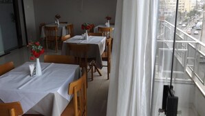 A dining area with tables set for four, each with a white tablecloth and a vase of red flowers. There are wooden chairs around the tables. The room has a large window with a view of a city street and buildings. - Hotel Nolasco (Macaé)