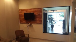 A living room with a flat-screen TV mounted on a wooden panel wall, a large window with a view of the outside, and a brown armchair. - Hotel Nolasco (Macaé)