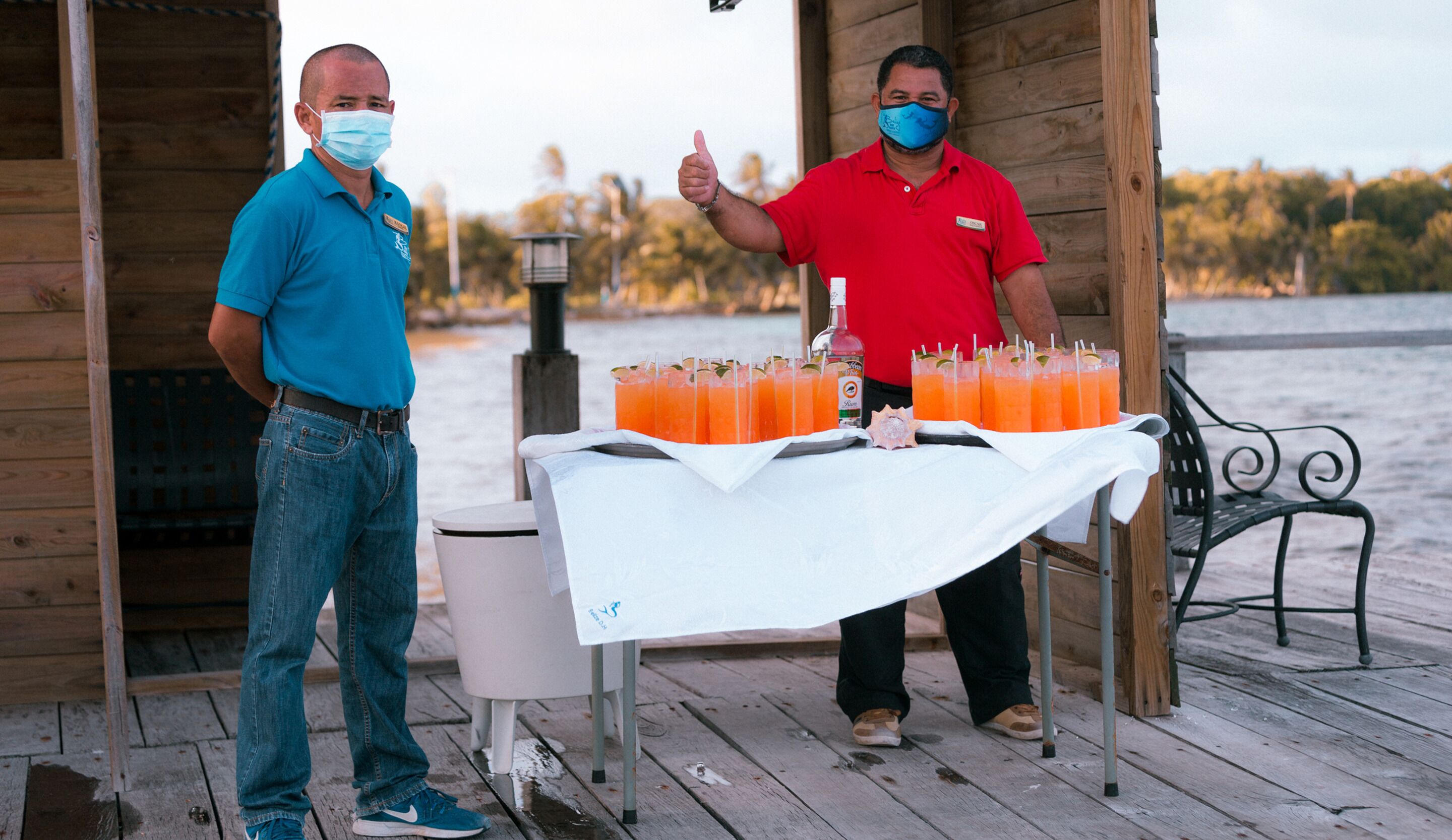 Two men standing by a table with drinks on a wooden deck by a body of water.