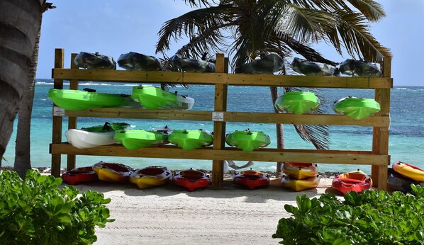 Un support en bois servant à ranger des kayaks, situé près de la plage, avec un palmier en arrière-plan et une vue sur l’océan.