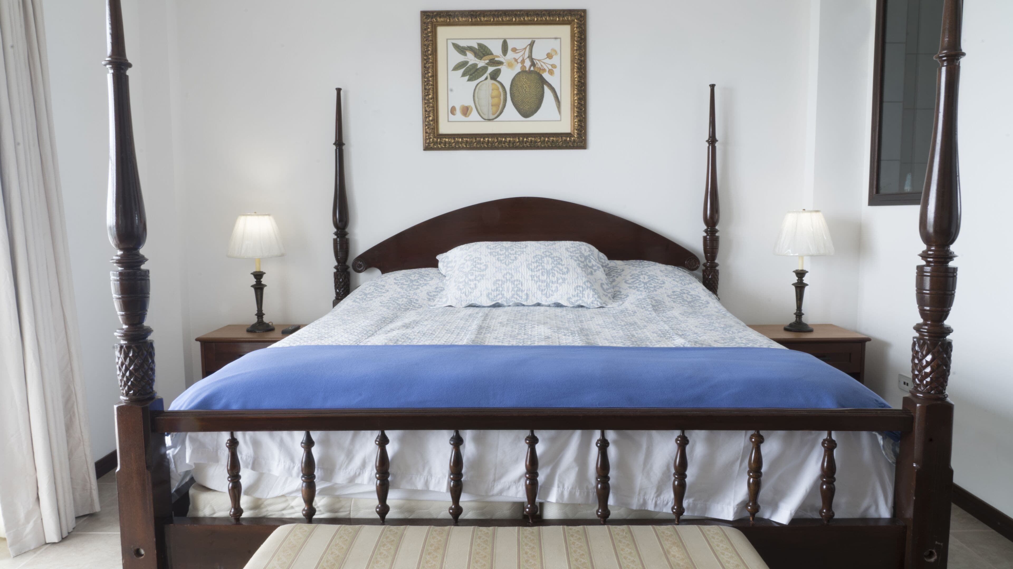 A four-poster bed with a blue bedspread and white pillows, flanked by two bedside tables with lamps, in a room with a framed botanical print on the wall.