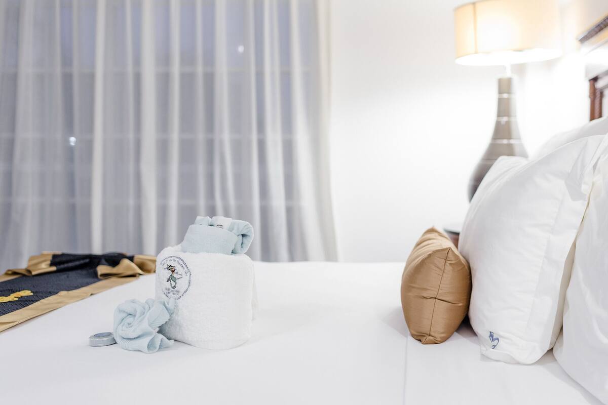 A neatly made bed with white linens, a brown pillow, and a folded towel with a logo.