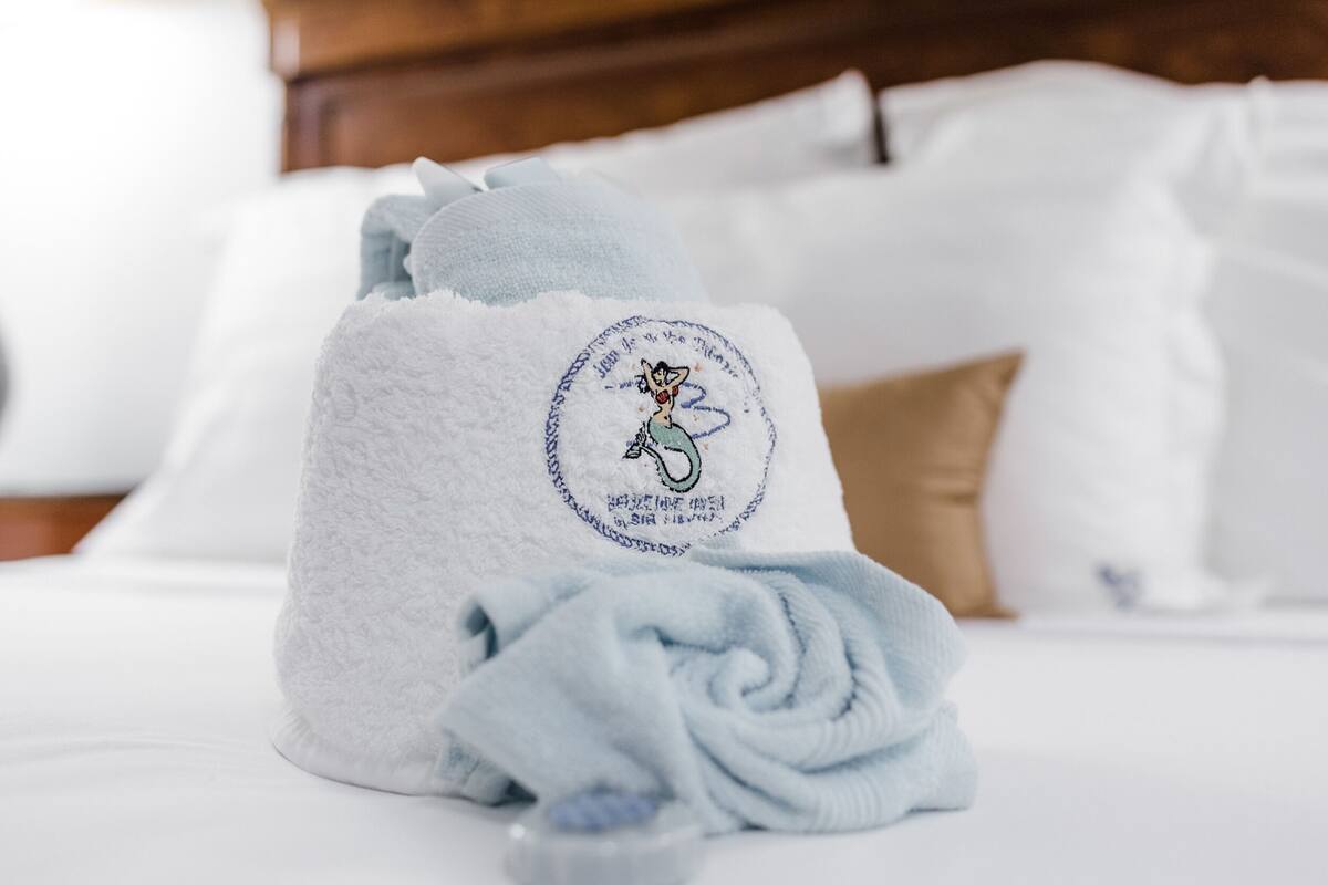 A neatly made bed with a towel featuring a mermaid design, white bedding, and a wooden headboard.