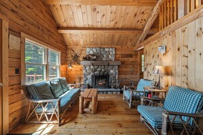 A log cabin interior with a stone fireplace, wooden furniture, and large windows.