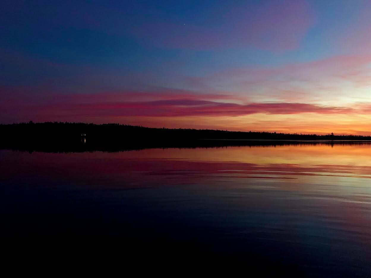 Un lac paisible au coucher du soleil, avec la silhouette d’arbres et une rive lointaine.