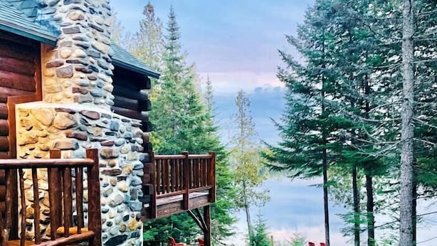 Une cabane dotée d’une cheminée en pierre, d’une terrasse en bois et d’un chemin de gravier menant à un lac bordé d’arbres.