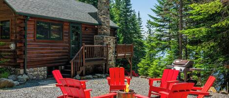A log cabin with a stone chimney, outdoor seating with red Adirondack chairs, and a fire pit.