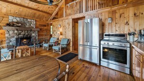 L’intérieur chaleureux d’un chalet avec une cheminée en pierre, un coin repas, une cuisine équipée d’un réfrigérateur et d’une cuisinière, et un plafond en bois.