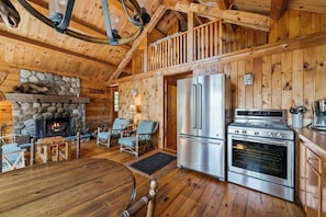 Un acogedor interior de cabaña con chimenea de piedra, un comedor, una cocina con refrigerador y estufa, y techo de madera.