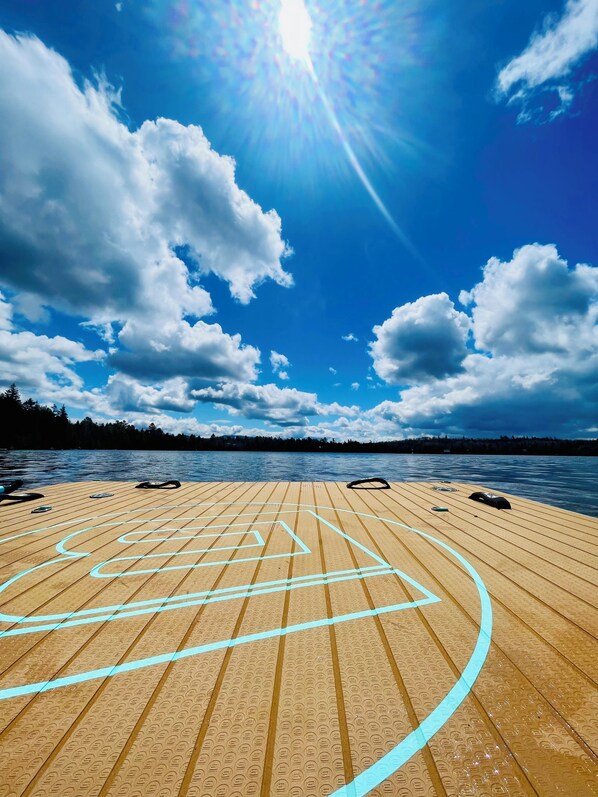 Una terraza de madera con líneas azules, una masa de agua y una orilla cubierta de bosque.