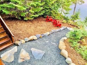 Un escalier en bois mène à une terrasse où se trouvent des fauteuils Adirondack rouges, et un chemin de gravier bordé de pierres.