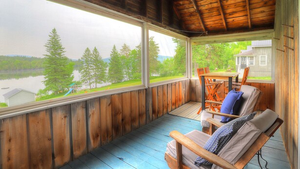A covered porch with a view of a lake and trees, wooden furniture, and a house in the background. - Hunter Cove Cabin #3: Shared Rangeley Lakefront, Dock Slip + Screened Porch (Rangeley)